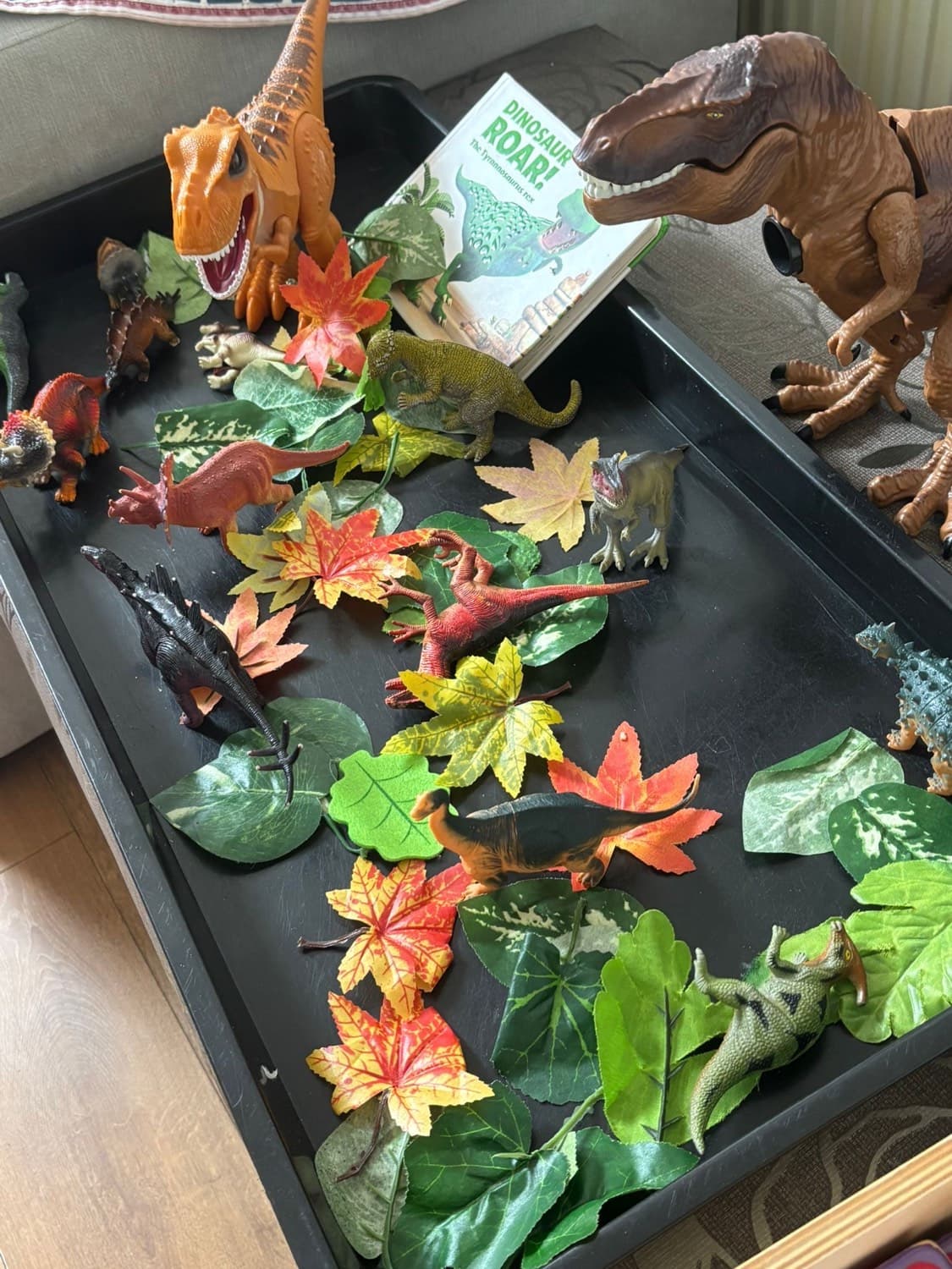 A small-world dinosaur play tray with toy dinosaurs, autumn leaves and a 'Dinosaur Roar!' picture book